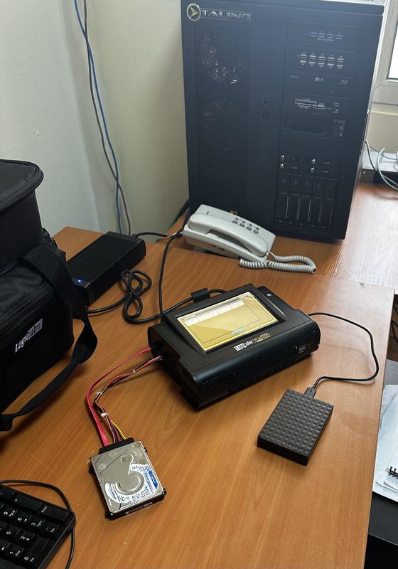 A Logicube forensic imager connected to a bare hard drive beside a forensic workstation